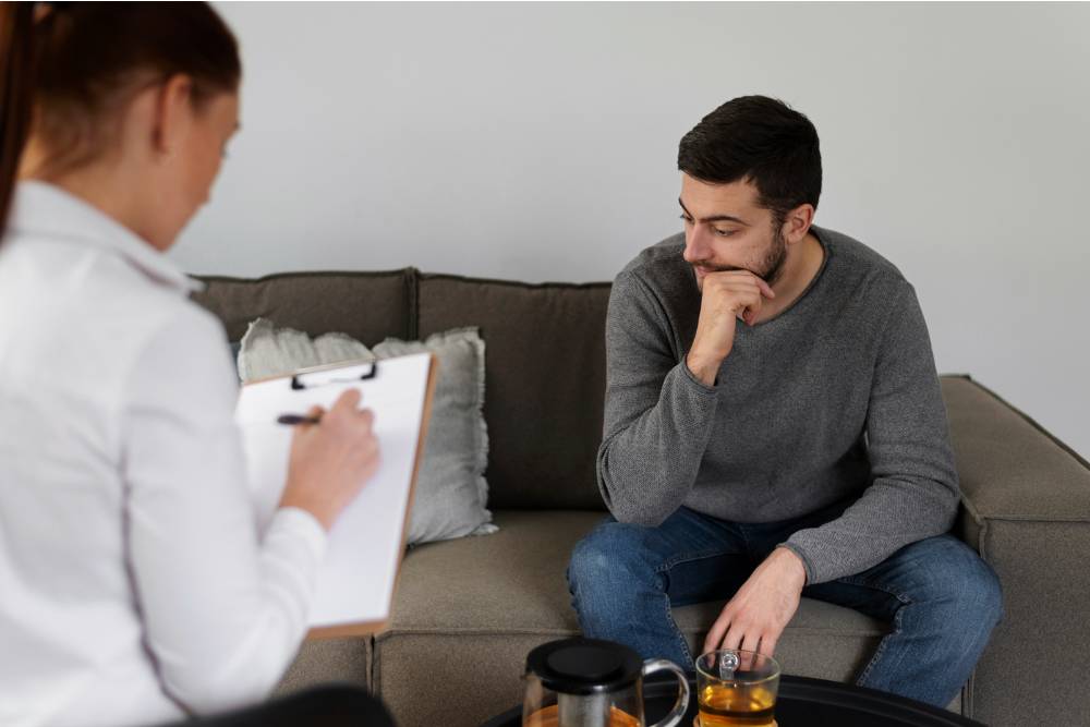 A therapist taking notes while a man sits pensively on a couch 
